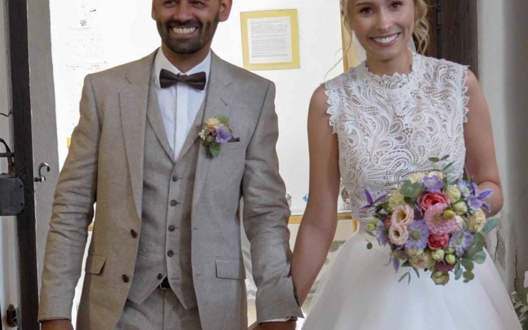 Ein Mann und eine Frau in Hochzeitskleidung schreiten gemeinsam den Gang entlang, umgeben von festlicher Atmosphäre.